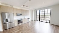 Located at 1 Main St in Miramar, FL, this efficiently designed single-floor unit offers a thoughtfully organized layout comprising a living room and a full bathroom. Light wood-tone flooring flows continuously throughout the space, complementing the clean white walls and creating a bright, cohesive aesthetic.

The open-concept living and kitchen area serves as the heart of the unit. The kitchen is outfitted with modern flat-panel cabinetry in a warm neutral finish, stainless steel appliances including a refrigerator, microwave, oven, and dishwasher, and a sleek countertop with an undermount sink. Track lighting overhead adds a contemporary touch and ensures ample illumination.

The living area is open and flexible, with clean lines and a minimalist design that maximizes the sense of space. A striking wall of floor-to-ceiling sliding glass doors with bold black grid-style framing leads to a private balcony, flooding the interior with natural light and offering expansive views of the surrounding Miramar landscape.

The bathroom features a glass-enclosed walk-in shower with a ceiling-mounted rain showerhead and polished chrome fixtures, set against crisp white tile walls — delivering a clean, spa-like feel. Overall, this unit presents a modern, functional living environment with stylish finishes throughout.