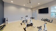 This single-floor fitness studio at 6255 Fernwood Dr. in Kannapolis, NC is purpose-built for cycling and group exercise. The space is designed with clean, modern finishes, anchored by striking light-toned herringbone-patterned flooring that lends a polished, contemporary feel throughout. The room is outfitted with multiple professional-grade spinning bikes, providing ample space for group cycling sessions. Recessed lighting and a ceiling fan ensure the space remains well-lit and comfortable during workouts. A large wall-mounted flat-screen television is prominently positioned, making it easy to follow along with on-screen fitness content from virtually anywhere in the room. A frosted-glass door with a transom window serves as the primary entry point, contributing to the studio's bright, open aesthetic while maintaining visual privacy. Multiple windows fitted with white blinds line the walls, inviting natural light and offering views of the surrounding outdoor environment. A bold navy blue accent wall adds a dynamic visual contrast to the otherwise neutral gray walls, energizing the space. Finishing touches such as white baseboards and a stainless steel sanitation station reflect thoughtful attention to both style and functionality, making this studio a well-equipped and inviting environment for fitness enthusiasts.