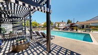 Located at 4022 Papillon Ter in Fremont, California, this outdoor community amenity space offers a well-appointed pool area designed for relaxation and recreation. The centerpiece is a generously sized swimming pool with clear, blue water, framed by a wide concrete deck that provides ample room for lounging and socializing.

A striking geometric pergola runs along one side of the pool, casting patterned shade over a row of padded chaise lounges — an ideal retreat from the California sun. On the opposite side of the deck, open sun loungers and umbrella-shaded seating arrangements with Adirondack-style chairs offer additional spots to unwind. String lights are strung overhead, adding a warm ambiance for evening gatherings.

The space also features a built-in outdoor kitchen equipped with a stainless steel grill and prep area, perfect for community barbecues and events. A ping pong table provides additional recreational options. The entire pool area is enclosed by a secure iron fence, with lush greenery, mature trees, and maintained lawns visible beyond the perimeter, lending a pleasant, park-like atmosphere. Under the abundant Fremont sunshine and with views of the surrounding hills, this outdoor space delivers a complete and inviting community leisure experience.