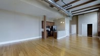 This single-floor loft space showcases a compelling blend of industrial character and modern functionality. The open-concept layout is anchored by warm hardwood floors that stretch throughout, creating a bright and cohesive feel across the living and kitchen areas. Soaring ceilings draw the eye upward to reveal exposed silver ductwork and original heavy timber beams — hallmark features of classic loft-style architecture that lend the space a raw, authentic industrial charm.

The kitchen is thoughtfully integrated into the open floor plan, centered around a structural wood post that adds architectural interest. Rich, medium-toned cabinetry provides ample storage and pairs well with granite countertops and a suite of stainless steel appliances, including a refrigerator with ice maker, electric range, and built-in microwave. A peninsula counter offers casual seating and separates the kitchen from the expansive living area without interrupting the open sightlines.

Pendant lighting above the kitchen adds a warm, inviting glow, complementing the natural light that fills the space. Additional rooms are accessible via interior doors visible along the perimeter, suggesting private spaces tucked beyond the main living area. The overall ambiance is one of urban sophistication — open, airy, and full of character.