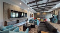 This well-appointed community clubroom delivers a dynamic, multi-use social environment designed for both relaxation and entertainment. The space is anchored by a striking media wall finished in weathered wood-toned panels, housing dual large flat-screen televisions above a built-in media console. Comfortable seating arrangements — including teal swivel chairs, a dark leather sofa, and a patterned area rug — create an inviting lounge atmosphere ideal for gathering and unwinding.

A built-in bookcase along one wall adds a warm, residential touch, stocked with books and decorative accents. The ceiling is a standout architectural feature, showcasing an exposed dark-stained beam coffered design that lends depth and character to the interior. Recessed lighting complements the natural light filtering in through multiple windows dressed with floor-length drapes.

Transitioning into the game zone, the space features two round poker-style tables surrounded by vibrant red, yellow, and teal chairs, alongside classic arcade game cabinets positioned near the windows. Colorful contemporary artwork punctuates the neutral walls, adding visual energy throughout. The cohesive patterned carpeting ties the entire space together, creating a seamless flow between the lounge and gaming areas. This clubroom is thoughtfully designed to encourage community interaction and leisure.