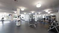 This fitness center offers a spacious, single-floor layout designed to accommodate a full range of workout activities. The space is finished with a durable dark rubber flooring, providing a practical and resilient surface suited for high-traffic athletic use. Smooth white columns punctuate the open floor plan, adding architectural structure while maintaining an airy, unobstructed feel throughout the room. The ceiling features recessed panels and evenly spaced circular flush-mount light fixtures that provide consistent, bright illumination across the entire space. Large windows along one wall allow natural light to filter in, enhancing the open and energizing atmosphere. Floor-to-ceiling mirrors line multiple walls, visually expanding the space and allowing users to monitor their form during workouts. The equipment layout is thoughtfully organized, with cardio machines — including treadmills and an elliptical — positioned near the windows, while a multi-station cable machine and weight benches occupy the central area. Exercise mats are also available for stretching or floor-based training. The overall design is clean and modern, with a white-and-grey color palette that conveys a professional, welcoming environment suited for a variety of fitness routines.