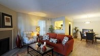 This single-floor residence offers a comfortable and well-organized layout designed for easy everyday living. The living area serves as the heart of the space, featuring light-toned walls, a textured ceiling, and warm artificial lighting that creates a cozy, welcoming atmosphere. The room comfortably accommodates a full seating arrangement, with light wood-style flooring that adds warmth and continuity underfoot. A standout architectural detail is the pass-through window connecting the living area to the kitchen, allowing for an open sightline and a sense of connectivity between the two spaces. The kitchen, visible through the pass-through, is well-lit with overhead fixtures and includes standard appliances, offering a functional cooking environment. Lush indoor plants are positioned throughout the living area, adding a natural, refreshing element to the interior. A hallway leading off the main living space suggests additional rooms beyond, contributing to the overall sense of a thoughtfully arranged floor plan. The combination of neutral wall colors, warm lighting, and an open, airy feel gives this space a practical yet inviting character suited to comfortable day-to-day living.