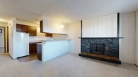 Located at 9310 E Montgomery Ave in Spokane Valley, WA, this single-floor unit offers a well-organized layout with a blend of classic and functional design elements throughout. The main living area is anchored by a striking floor-to-ceiling stone fireplace with a glass-front insert and a wooden hearth, set against white vertical-panel accent walls that add architectural character. A pendant light fixture provides warm overhead illumination, and a textured ceiling runs throughout the space. The kitchen features warm wood cabinetry, laminate countertops, wood-look flooring, a white electric range, dishwasher, and a double sink. A breakfast bar peninsula opens toward the living area, creating a connected, efficient flow. A separate bedroom offers generous storage with two sets of bifold closet doors and is outfitted with a ceiling fan and a sliding glass door that brings in natural light and outdoor access. Baseboard heaters provide supplemental warmth in the carpeted rooms. The full bathroom includes a tub-shower combo, a wood-cabinet vanity, and wood-look flooring. The entry foyer features durable dark tile flooring, a front-facing window with blinds, and a solid front door, providing a clean and practical welcome to the space.