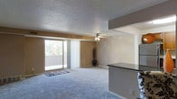 This single-floor residence showcases an open-concept layout that connects the living area to the kitchen space. The living room is finished with plush wall-to-wall carpeting and warm neutral-toned walls, creating a comfortable and inviting atmosphere. A ceiling fan with integrated lighting is centrally mounted on the textured popcorn ceiling, providing both airflow and ambient light to the space. A full-width sliding glass door along the back wall opens to an outdoor balcony, allowing natural light to flow generously into the interior and offering views of surrounding greenery. Vertical blinds on the sliding door provide privacy and light control as needed. Adjacent to the living area, a raised counter with a dark laminate surface serves as a functional partition between the living space and the kitchen, offering a convenient breakfast bar or casual dining area. The overall design is practical and straightforward, with the open flow between rooms enhancing the sense of space throughout the single-floor layout.