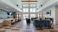 This expansive single-floor amenity space is defined by its striking architectural design and sophisticated interior finishes. The central great room commands attention with soaring double-height ceilings accented by exposed wood beams and a dramatic floor-to-ceiling window arrangement that floods the space with natural light and offers direct views of an outdoor pool and surrounding landscaping. A statement chandelier hangs at the heart of the room, complemented by gold cone-shaped wall sconces that add warmth and character throughout.

The multi-toned tile flooring features a distinctive geometric patchwork pattern that flows seamlessly across the entire space, lending visual interest and cohesion. The open-plan layout accommodates distinct seating areas furnished with plush navy and teal upholstered sofas and armchairs, anchored by patterned area rugs. A fireplace serves as a focal point in one lounging area, adding comfort and ambiance.

A formal dining area with a long wood table and upholstered seating sits adjacent to the main lounge, defined by bold decorative wallcoverings and pendant lighting overhead. The entry corridor features rich wood-toned double doors flanked by framed artwork, creating a refined arrival experience. Large glass partitions and French-style doors connect interior zones, enhancing openness while maintaining functional separation throughout this well-appointed communal space.