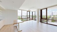 This well-appointed 5-bedroom, 2-bathroom unit is designed with a clean, contemporary aesthetic throughout its single floor. Light hardwood flooring runs continuously across the space, complementing the crisp white walls and ceilings that create a bright, airy atmosphere.

The open-concept living area is a standout feature, anchored by dramatic floor-to-ceiling corner windows that flood the space with natural light and frame expansive views of the surrounding cityscape, including a prominent skyline visible in the distance. The integrated kitchen is outfitted with stainless steel appliances, light wood cabinetry, and a gas range. A private balcony accessible from the living area extends the city views outdoors with a full railing for safety.

The five bedrooms maintain the home's clean lines and light palette, with several featuring large floor-to-ceiling windows that continue the panoramic urban views. The primary bedroom is generously proportioned and benefits from ample natural light. A dedicated walk-in closet with built-in shelving and hanging rods provides substantial storage.

The bathrooms are finished in white tile, with one featuring a glass-enclosed walk-in shower with a recessed niche and chrome fixtures. Two hallways connect the layout efficiently, and an in-unit laundry area adds everyday convenience. Smart home technology, including a digital thermostat, is also present throughout.