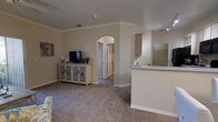This single-floor, 2-bedroom, 2-bathroom residence is thoughtfully designed to maximize comfort and functionality. The open-concept living area features plush carpeting, neutral warm-toned walls, and crown molding that lends a polished, refined look throughout. Elegant arched doorways serve as architectural focal points, adding character and a sense of flow between spaces.

The living room is generously proportioned, providing ample room for furnishings and everyday living. It connects seamlessly to the kitchen area, which features a raised breakfast bar peninsula — ideal for casual dining or additional workspace. The kitchen is equipped with light-toned cabinetry and a functional layout designed for efficient use of space.

Crown molding detailing continues throughout the common areas, complementing the clean, neutral color palette that gives the home a bright and welcoming ambiance. Ceiling vents are integrated discreetly, ensuring consistent climate control. The arched hallway leads to the private bedroom and bathroom areas, offering a clear separation between living and sleeping quarters.

Overall, the residence presents a cohesive, comfortable interior with classic architectural detailing, practical design, and a neutral aesthetic that provides a versatile backdrop for a variety of décor styles.