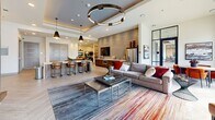 Located at 1703 Rockhill Rd in McKinney, TX, this single-floor community amenity space offers a generously sized, open-concept layout designed for both relaxation and social gathering. The interior showcases a sophisticated contemporary aesthetic, anchored by herringbone-patterned light wood tile flooring that flows seamlessly throughout.

A dramatic stone-clad feature wall serves as the focal point of the lounge area, housing a wall-mounted flat-screen TV above a sleek linear fireplace with a glass ember bed. Comfortable sectional seating, a marble-top coffee table, and a large area rug define this welcoming lounge zone. Overhead, a bold circular chandelier adds a striking architectural element.

The space transitions fluidly into a kitchenette and bar area, complete with a white island counter, bar stools, pendant lighting, and stainless steel appliances. A billiards table occupies a dedicated game area nearby, ideal for recreational use.

A dining nook bathed in natural light features floor-to-ceiling glass walls and doors opening to an outdoor area with pool views. Sculptural pendant fixtures and curved wooden chairs lend a refined, boutique feel to this corner. Throughout, tray ceilings with recessed lighting and large windows ensure the space remains bright, airy, and inviting.