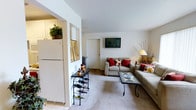 This two-floor residence offers a thoughtfully arranged layout designed for comfortable everyday living. The living room serves as the heart of the home, featuring soft carpeted flooring, neutral white walls, and a gently vaulted ceiling that adds a sense of openness to the space. A wall-mounted flat-screen television anchors one side of the room, while plush sofas adorned with decorative red and floral accent pillows create a warm and inviting seating arrangement. A mosaic-tiled coffee table and a wrought iron wine rack add charming decorative touches that lend character to the space. An elegant floor lamp provides warm ambient lighting, complementing the overall cozy atmosphere. The living area connects seamlessly to the kitchen through an open archway, where white cabinetry and a full-size refrigerator are visible, hinting at a practical and well-equipped culinary space. Lush potted greenery displayed atop the kitchen cabinets adds a refreshing natural element throughout. The transition between the tiled kitchen flooring and the carpeted living area clearly delineates the two functional zones while maintaining a cohesive, welcoming feel across the home's main level.