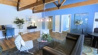This single-floor space makes a strong architectural impression with its exposed wood beam ceiling, supported by black metal hardware that adds an industrial contrast to the warm timber. A modern chandelier with Edison-style bulbs hangs as a central focal point, illuminating the open layout below. The flooring throughout is rich hardwood, grounding the space with warmth and continuity.

Bold blue accent walls energize the interior, complementing the crisp white walls and trim for a balanced, contemporary aesthetic. The main living area is generously proportioned, furnished with dark leather seating arranged around a glass-top coffee table atop a textured area rug, conveying a comfortable yet stylish atmosphere. Chevron-patterned accent chairs add a playful geometric element to the seating arrangement.

A sleek black console table against the wall provides functional display space, accented by a table lamp and decorative items. Multiple white doors lead to additional rooms or spaces, one of which reveals a glimpse of a secondary room with its own natural light. The overall ambiance is open, modern, and well-appointed, blending rustic architectural elements with contemporary furnishings.