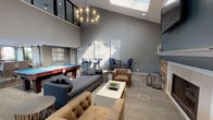 The Raintree Apartments community clubhouse is a thoughtfully designed gathering space that blends comfort and modern style throughout. The main lounge area commands attention with its soaring vaulted ceilings, a large skylight that floods the room with natural light, and a striking statement chandelier. A wood-accented fireplace wall serves as a dramatic focal point, featuring reclaimed-style planking, a flat-screen TV above the mantle, and rich navy accent walls that lend a sophisticated ambiance. Comfortable tufted leather armchairs and a plush sectional sofa invite relaxation, while a full-size billiards table adds a recreational element to the space. Decorative mandala artwork adorns the walls alongside bistro-height pub tables and upholstered bar stools, creating a casual social atmosphere. A separate lounge nook features bold two-tone walls with a patterned area rug, ideal for quieter, more intimate seating. The fully equipped kitchen is a standout feature, outfitted with stainless steel appliances, granite countertops, a large prep island with bar seating, wood-look tile flooring, and pendant lighting. Expansive floor-to-ceiling windows in the kitchen area provide direct views of the outdoor pool and surrounding landscaping, seamlessly connecting the interior with the resort-style amenities outside.