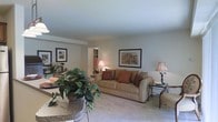 Located at 2901 Crossfork Drive in Wilmington, Delaware, this single-story residence offers a practical and inviting layout designed for comfortable everyday living. The living area serves as the heart of the home, featuring plush wall-to-wall carpeting that adds warmth underfoot and contributes to the space's cozy atmosphere. Neutral-toned walls provide a versatile backdrop, complemented by soft, ambient lighting from elegant floor and table lamps that create a relaxed and welcoming ambiance. The living room accommodates generous seating, as evidenced by the large sofa accented with decorative throw pillows in warm earth tones. Framed artwork on the walls adds a refined, homey character to the space. A glass-top side table provides a functional accent piece alongside the seating area. Adjacent to the living room, a passage leads to what appears to be a closeted bedroom or additional room, suggesting a well-connected interior flow. The open sightlines between the living and dining areas, along with lush indoor greenery visible in the foreground, enhance the sense of spaciousness and bring a natural element into the home's overall aesthetic.