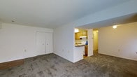 This single-floor unit offers a straightforward and functional layout designed for comfortable everyday living. The main living area is finished with wall-to-wall carpeting in a neutral tone, providing a warm and quiet underfoot feel throughout the space. A set of double bi-fold closet doors along one wall offers convenient built-in storage, keeping the living area clean and uncluttered.

A half-wall partition separates the living space from the kitchen, creating a sense of openness while maintaining a distinct cooking zone. The kitchen features warm honey-toned wood cabinetry, a white range, and an overhead range hood, providing all the essentials for meal preparation. The layout allows for easy flow between the kitchen and the adjacent hallway, which leads further into the unit.

The hallway connects to additional areas of the home, hinted at by the warm lighting visible beyond the kitchen passthrough. The overall color palette throughout the unit is light and neutral, with white walls that help maximize the sense of brightness in the space. The single-floor plan ensures seamless, step-free accessibility throughout. This unit presents a practical and well-organized living environment suited to a variety of lifestyles.