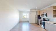 This single-floor unit offers a clean, contemporary design throughout. The open-concept layout seamlessly connects the living area and kitchen, creating a bright and functional shared space. Light wood-tone laminate flooring runs continuously throughout, contributing to a cohesive, modern aesthetic.

The kitchen is well-appointed with light wood cabinetry, open shelving, and white subway tile backsplash. Stainless steel appliances include a range, microwave, dishwasher, and refrigerator, alongside a undermount sink with light-toned countertops. Recessed lighting illuminates the kitchen efficiently.

The living area is open and versatile, featuring crisp white walls, multiple electrical outlets, and a window that allows natural light and outdoor views of the surrounding neighborhood. The space is generously proportioned, offering flexibility for various furniture arrangements.

The bathroom maintains the same modern aesthetic, featuring a wood-toned vanity with an undermount sink, white countertop, a large mirror, and a gold-toned wall sconce above. Light wood flooring continues here, complementing the clean white walls. Overall, the unit presents a bright, minimalist character with consistent finishes throughout.