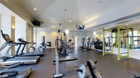 This fitness center offers a comprehensive workout environment designed to accommodate a variety of exercise needs. The space features durable dark flooring throughout, providing a functional and professional aesthetic well-suited to an active setting. Recessed lighting illuminates the room evenly, complemented by natural light filtering in through upper clerestory windows that lend an open, airy feel to the interior. The ceiling is finished with subtle architectural detailing, adding a refined touch to the otherwise utilitarian space. Multiple flat-screen televisions are mounted at strategic points, keeping members entertained during workouts. The facility is generously stocked with cardio equipment, including treadmills, elliptical trainers, and stationary bikes. A dedicated strength training area features a multi-function cable machine and a power rack with weight plates and barbells for free weight exercises. A full-length mirror along one wall enhances the sense of spaciousness while allowing members to monitor their form. The layout is thoughtfully organized to allow for comfortable movement between stations. An exit door is visible at the rear, and the overall condition of the facility reflects a well-maintained, professionally managed space.