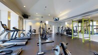 This fitness center offers a comprehensive workout environment designed to accommodate a variety of exercise needs. The space features durable dark flooring throughout, providing a functional and professional aesthetic well-suited to an active setting. Recessed lighting illuminates the room evenly, complemented by natural light filtering in through upper clerestory windows that lend an open, airy feel to the interior. The ceiling is finished with subtle architectural detailing, adding a refined touch to the otherwise utilitarian space. Multiple flat-screen televisions are mounted at strategic points, keeping members entertained during workouts. The facility is generously stocked with cardio equipment, including treadmills, elliptical trainers, and stationary bikes. A dedicated strength training area features a multi-function cable machine and a power rack with weight plates and barbells for free weight exercises. A full-length mirror along one wall enhances the sense of spaciousness while allowing members to monitor their form. The layout is thoughtfully organized to allow for comfortable movement between stations. An exit door is visible at the rear, and the overall condition of the facility reflects a well-maintained, professionally managed space.