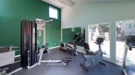 This fitness center offers a functional and well-lit workout environment designed to support a variety of exercise routines. The space features soaring vaulted ceilings that lend an open, airy feel, complemented by large windows and glass doors that flood the room with natural light and provide views of an outdoor pool area. The bold green accent walls contrast with the light mint-painted surfaces, creating a vibrant and energizing atmosphere throughout.

The workout area is equipped with a range of fitness equipment, including a treadmill, two elliptical cross-trainers, a stationary recumbent bike, and a multi-station cable weight machine with a dedicated flat bench for strength training. A wall-mounted split-unit air conditioning system ensures a comfortable climate during workouts. The dark rubberized flooring provides durability and traction underfoot, making the space both practical and safe for exercise.

Full-length mirrors are mounted along one wall, allowing users to monitor their form while training and visually expanding the sense of space. Green-framed entry doors provide direct access to the outdoor grounds and pool area, seamlessly connecting the indoor fitness space with the exterior amenities. Overall, this fitness center is a bright, well-appointed space suited for both cardio and strength training.