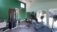 This fitness center offers a functional and well-lit workout environment designed to support a variety of exercise routines. The space features soaring vaulted ceilings that lend an open, airy feel, complemented by large windows and glass doors that flood the room with natural light and provide views of an outdoor pool area. The bold green accent walls contrast with the light mint-painted surfaces, creating a vibrant and energizing atmosphere throughout.

The workout area is equipped with a range of fitness equipment, including a treadmill, two elliptical cross-trainers, a stationary recumbent bike, and a multi-station cable weight machine with a dedicated flat bench for strength training. A wall-mounted split-unit air conditioning system ensures a comfortable climate during workouts. The dark rubberized flooring provides durability and traction underfoot, making the space both practical and safe for exercise.

Full-length mirrors are mounted along one wall, allowing users to monitor their form while training and visually expanding the sense of space. Green-framed entry doors provide direct access to the outdoor grounds and pool area, seamlessly connecting the indoor fitness space with the exterior amenities. Overall, this fitness center is a bright, well-appointed space suited for both cardio and strength training.
