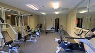 This single-floor fitness room offers a practical and functional workout space designed to accommodate a range of exercise needs. The room features a textured ceiling with recessed fluorescent lighting panels that provide bright, even illumination throughout the space. Neutral beige walls and speckled grey carpeted flooring create a clean, no-frills environment suited for focused training sessions.

The layout is thoughtfully organized to maximize the available floor space. Cardio equipment includes a Cybex rowing machine and a stationary recumbent bike, while strength training options feature a multi-function cable machine, a power rack, and multiple weight benches. A medicine ball and additional free-standing equipment further round out the selection, supporting both resistance and functional training routines.

A large mirror on one wall enhances the sense of space and allows users to monitor their form during exercise. Ventilation is provided via ceiling vents, contributing to a comfortable environment during workouts. Two doorways provide convenient access points to the room, and the overall layout allows for adequate circulation between machines.