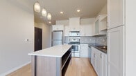 This thoughtfully designed single-floor unit offers a clean, contemporary aesthetic throughout. The kitchen serves as a central highlight, featuring crisp white shaker cabinetry, stainless steel appliances including a range, microwave, refrigerator, and dishwasher, complemented by a neutral tile backsplash and dark countertops. A large central island with a white quartz surface and wood-toned base provides additional workspace and storage, while pendant lighting adds a stylish finishing touch overhead. Light hardwood flooring runs throughout the kitchen and main living areas, contributing to a bright and cohesive feel. The living spaces benefit from oversized windows fitted with horizontal blinds, allowing abundant natural light and outward views of the surrounding neighborhood. Bedroom spaces are finished with plush carpeting and large windows that enhance the sense of openness. The bathroom is clean and functional, featuring a combined tub and shower, a vanity with storage, and consistent light flooring. A dedicated laundry area houses a full-sized top-load washer and dryer, adding everyday convenience. The overall design is modern and understated, with a neutral color palette that creates a calm, versatile living environment throughout.