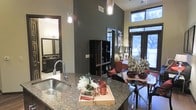 This single-floor space showcases a thoughtfully designed open-concept layout that effortlessly connects the kitchen, dining, and living areas into one cohesive environment. The kitchen is a standout feature, boasting rich granite countertops, a deep stainless steel sink, and sleek pendant lighting that hangs dramatically from the soaring ceilings above — adding both function and visual interest. The dark, bold wall tones lend a sophisticated and contemporary character throughout, complementing the dark-trimmed doorways and cabinetry. Large floor-to-ceiling windows along the exterior wall flood the space with natural light, offering views of the outdoors and creating an airy, open ambiance despite the intimate scale. A dining area sits just off the kitchen, providing a natural transition into the living space beyond. A stylish open shelving unit and tasteful décor accents add personality and depth to the interior. A bathroom is also visible, featuring a well-appointed vanity with dark fixtures that maintain the cohesive modern aesthetic. Overall, the space strikes a balance between bold design choices and welcoming warmth, making it an attractive and highly functional living environment.