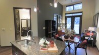 This single-floor space showcases a thoughtfully designed open-concept layout that effortlessly connects the kitchen, dining, and living areas into one cohesive environment. The kitchen is a standout feature, boasting rich granite countertops, a deep stainless steel sink, and sleek pendant lighting that hangs dramatically from the soaring ceilings above — adding both function and visual interest. The dark, bold wall tones lend a sophisticated and contemporary character throughout, complementing the dark-trimmed doorways and cabinetry. Large floor-to-ceiling windows along the exterior wall flood the space with natural light, offering views of the outdoors and creating an airy, open ambiance despite the intimate scale. A dining area sits just off the kitchen, providing a natural transition into the living space beyond. A stylish open shelving unit and tasteful décor accents add personality and depth to the interior. A bathroom is also visible, featuring a well-appointed vanity with dark fixtures that maintain the cohesive modern aesthetic. Overall, the space strikes a balance between bold design choices and welcoming warmth, making it an attractive and highly functional living environment.