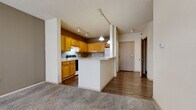 This single-floor residential unit offers a well-organized layout that balances open communal spaces with private rooms. The kitchen is a standout feature, showcasing warm honey-toned wood cabinetry that extends to the ceiling, providing ample storage throughout. The kitchen is equipped with a full suite of white appliances, including a refrigerator, dishwasher, and electric range, all set atop durable hardwood-style flooring. A peninsula with a light countertop extends into the living area, creating a natural divide between the kitchen and the adjoining space while offering additional seating or prep area. Track lighting runs throughout the main living areas, providing flexible, adjustable illumination. The living room is generously sized and finished with plush carpeting in a neutral gray tone, offering a comfortable, versatile space. Multiple bedroom areas echo the same soft carpet and neutral wall colors, with one bedroom featuring a window that allows natural light to filter in. The bathroom is clean and functional, featuring a wide vanity with a single basin sink, warm wood cabinetry below, a large mirror, and a tub-shower combination with grab bars. Throughout the unit, a consistent neutral color palette of whites, creams, and warm grays creates a cohesive and inviting ambiance.
