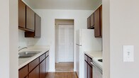 This single-floor unit offers a practical and well-organized layout designed to maximize functionality throughout. The galley-style kitchen features rich dark wood cabinetry lining both walls, light countertops, hardwood flooring, and white appliances including a refrigerator and stovetop. A doorway at the end of the kitchen leads directly into the main hallway, creating a smooth flow through the unit.

The living area is generously sized, filled with natural light from a large sliding glass door that opens to an outdoor view, complemented by an additional window with vertical blinds. Neutral carpet and light-toned walls give the space an open, airy feel.

The hallway connecting the main living spaces is lined with louvered bifold closet doors, offering ample built-in storage. A ceiling light fixture illuminates the passage, which leads to the bathroom and bedrooms.

The bathroom features classic small-tile flooring in a warm tone, a dark wood vanity with a wide countertop, and a full mirror, providing both style and practicality.

The bedrooms are carpeted and well-proportioned, each featuring neutral walls, bifold closet doors with rod storage, and windows dressed with vertical blinds that invite natural light. The overall ambiance of the unit is clean, neutral, and move-in ready.