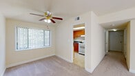 This single-floor residence offers a well-organized layout with multiple rooms designed for comfortable living. The expansive living area is finished with plush wall-to-wall carpeting and features ceiling fans with integrated lighting throughout. Large windows dressed with vertical blinds allow generous amounts of natural light to fill each room, creating a bright and airy atmosphere. The layout, as visible from the dollhouse view, includes distinct living and sleeping areas connected by a central corridor, providing a functional flow throughout the unit. The kitchen is neatly appointed with warm oak-toned cabinetry extending to the ceiling for ample storage, dark countertops with an integrated sink, a gas range, and a full-size refrigerator. Light tile flooring and a window with vertical blinds complete the kitchen's practical design. The bathroom includes a full-size soaking tub with white tile surround, a vanity with wood cabinetry, and a window that brings in natural light and outdoor greenery views. Bedroom spaces are carpeted and generously sized, each equipped with ceiling fans and windows overlooking lush outdoor surroundings. The overall color palette of warm cream tones and white trim gives the space a clean, neutral, and inviting character throughout.