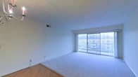 This single-floor residence offers a clean, open layout designed to maximize comfort and natural light. The main living area is a standout feature, anchored by a wide wall of floor-to-ceiling sliding glass doors fitted with vertical blinds. These doors flood the space with daylight and provide direct views of the surrounding neighborhood, including adjacent buildings and mature trees — giving the room a bright, airy character throughout the day.

The living area is finished with wall-to-wall plush carpeting in a neutral tone, creating a soft and welcoming atmosphere. The smooth white walls and clean ceiling with a centrally placed ventilation register contribute to the space's minimalist and well-maintained aesthetic. A transition to hardwood-style flooring is visible at the entry point of the room, adding a subtle contrast in materials and defining the boundary between living and entry zones.

The overall design is simple and functional, with a neutral color palette throughout that makes the space highly adaptable to a variety of furnishing styles. The abundance of natural light from the expansive glazed wall ensures the interior feels open and connected to the outdoors, while the elevated positioning of the unit enhances the sense of space and provides clear sightlines to the exterior environment.