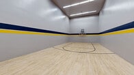 Located at 240 S Monaco Street Parkway in Denver, Colorado, this indoor sports facility offers a well-appointed space designed for both basketball and racquetball. The facility spans a generous single-floor footprint divided into two distinct court areas connected by a hallway, providing dedicated zones for each sport. Both courts feature smooth, light-toned hardwood flooring with clearly marked black court lines, offering excellent traction and a professional playing surface. A bold decorative stripe of navy blue and yellow runs along the perimeter walls of each court, adding a sporty, energetic visual identity to the space. The basketball court is equipped with a wall-mounted, clear-backboard hoop and features standard court markings including the three-point arc and key, making it suitable for organized play or casual games. The racquetball court offers a clean, enclosed playing environment with tall ceilings and smooth, unobstructed walls — ideal for competitive rallies. Overhead fluorescent lighting illuminates both courts evenly, ensuring consistent visibility throughout. The facility's straightforward, functional design prioritizes athletic performance while maintaining a clean and well-maintained aesthetic throughout.