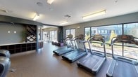 Located at 70 Upper Rock Circle in Rockville, Maryland, this comprehensive fitness center is designed to support a full range of workout routines in a modern, well-appointed environment. The space features durable rubber flooring throughout, smooth white ceilings with bright overhead lighting, and neutral gray walls that create a clean, focused atmosphere.

The cardio zone is equipped with multiple treadmills and Cybex elliptical machines, positioned along large windows that flood the area with natural light and offer views of the surrounding Upper Rock Circle development. A dedicated cycling alcove provides a more enclosed setting for stationary bike workouts.

The strength training area is outfitted with Hoist cable machines, plate-loaded equipment, and adjustable benches, complemented by a full rack of dumbbells displayed in front of wall-mounted mirrors that enhance the sense of space. A flat-screen television is mounted nearby for entertainment during workouts.

Structural columns are integrated throughout the floor plan, contributing to the architectural character of the space. Additional amenities include a wall-mounted equipment organizer and decorative shelving, adding both functionality and visual interest. The generous layout ensures ample room between stations, allowing for comfortable, unobstructed movement throughout the entire fitness facility.