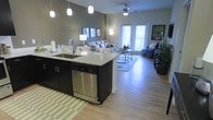 This single-floor space showcases an open-concept layout that seamlessly integrates the kitchen and living areas into one cohesive environment. The kitchen is anchored by a central island finished with light granite countertops and dark cabinetry, offering both prep space and a casual dining area. A stainless steel dishwasher and an undermount sink are built into the island, complemented by pendant lighting that hangs overhead. The transition into the living area is smooth and uninterrupted, with continuous hardwood-style flooring extending throughout. Sage-green accent walls provide a calming, neutral backdrop, while a ceiling fan overhead ensures comfort year-round. Natural light fills the space through a set of glass French doors, creating a bright and airy atmosphere. The living area comfortably accommodates a full seating arrangement, with room for accent furniture and décor. Overall, the space conveys a modern yet welcoming ambiance, designed for both everyday functionality and relaxed living.