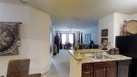 This single-floor residence offers an open-concept design that creates a seamless flow between the kitchen, dining, and living spaces. The kitchen is a functional centerpiece, featuring rich dark wood cabinetry, a double stainless steel sink, and a granite-topped breakfast bar with a gracefully curved edge that provides additional seating while maintaining an open sightline into the rest of the home. The cabinetry's deep tone contrasts elegantly with the light granite countertops, creating a balanced and refined aesthetic. Beyond the kitchen, the living area is generously proportioned, bathed in natural light from large windows framed by dark curtains that offer both style and privacy. Plush, light-toned carpeting extends throughout the living space, adding warmth and comfort underfoot. A ceiling fan with light fixture serves as a practical overhead fixture while contributing to the room's relaxed ambiance. The neutral wall tones provide a versatile backdrop for décor, and artwork and decorative accents lend a personal, curated feel to the space. Recessed lighting and built-in ventilation further enhance the home's modern comfort and functionality throughout.