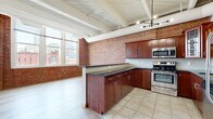 Located at 1300 West 9th Street in Cleveland's dynamic Tremont/near-West Side neighborhood, this two-bedroom, two-bathroom loft blends industrial character with modern comfort. The open-concept kitchen and living area serves as the heart of the space, showcasing exposed red brick walls, white-painted wood beam ceilings with visible ductwork, and large windows that flood the room with natural light and frame impressive views of the Cleveland skyline. The kitchen features rich cherry wood cabinetry, dark granite countertops, stainless steel appliances, and tile flooring that transitions into light hardwood in the living area. The primary bedroom continues the loft aesthetic with exposed beamed ceilings, a clerestory interior window, and light wood-tone flooring. The second bedroom offers carpeted floors, built-in closet storage with wood-panel doors, and its own stretch of exposed brick. Both bathrooms are finished with tile flooring, dark granite vanity tops, and matching cabinetry. Hallways connect each area efficiently, and a dedicated laundry space with an in-unit stacked washer and dryer adds everyday convenience. This space delivers an authentic urban loft experience with thoughtful finishes throughout.