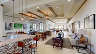This generously sized single-floor community clubhouse spans an expansive open footprint designed to accommodate a variety of social and recreational activities. The main gathering area showcases soaring ceilings adorned with decorative wood beam accents and recessed lighting complemented by stylish pendant fixtures. Large arched windows and glass panel doors flood the interior with natural light while offering views of the surrounding outdoor patio areas. Light-toned ceramic tile flooring runs throughout the primary spaces, creating a clean, cohesive aesthetic.

The open-plan layout seamlessly connects multiple lounge zones furnished with contemporary sofas, accent chairs, and coffee tables arranged for both casual conversation and relaxation. A granite-topped bar counter with barstool seating serves as a central gathering point. The dedicated game room features a full-size pool table, a foosball table, wall-mounted televisions, and bar-height seating along a tiled ledge — ideal for social recreation.

A separate area with wood-look plank flooring houses a business center workstation and additional seating with wall-mounted artwork. French doors connect transitional spaces, maintaining an open yet defined flow throughout. Warm wood trim detailing, curated artwork, and thoughtful furnishings throughout give the clubhouse a welcoming, upscale ambiance suited for community gatherings.