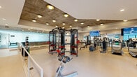 This modern fitness center offers a comprehensive workout environment spread across a single, open-plan floor. The space is defined by a striking exposed concrete ceiling inset with warm recessed lighting, creating an industrial yet inviting atmosphere that contrasts with the clean, light-toned flooring throughout.

The cardio zone is well-appointed with multiple treadmills, elliptical trainers, and rowing machines, all oriented toward large wall-mounted flat-screen televisions. Full-length mirrors along the walls visually expand the space and allow users to monitor their form during workouts.

At the heart of the fitness center stands a Life Fitness Synrgy360 functional training system, accompanied by kettlebells, medicine balls, and a variety of free weights and benches, catering to strength and functional training routines. A dedicated stretch and flexibility area features stability balls and open floor space, positioned near large windows that frame views of the surrounding outdoor courtyard, filling the area with natural light.

A vivid, large-scale athletic mural adorns one whitewashed brick accent wall, adding an energetic and motivational focal point to the space. A storage room provides additional utility. The overall design balances high-performance functionality with a polished, contemporary aesthetic suited for a wide range of fitness activities.