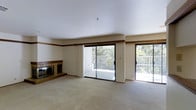 This single-floor residence offers a bright, open living space designed for both comfort and functionality. The main living area is generously sized, finished with wall-to-wall carpeting that adds warmth underfoot. A tiled fireplace with a raised hearth serves as a focal point on one wall, providing a classic and inviting centerpiece to the room. A warm wood trim accent rail runs along the upper portion of the walls, adding a touch of architectural character to the otherwise clean, neutral interior. Natural light flows abundantly into the space through multiple large sliding glass doors, which open directly onto a spacious outdoor deck. The deck extends the living area outward and is framed by mature trees and lush greenery, creating a serene, nature-connected atmosphere. The smooth white ceilings enhance the sense of openness throughout. The layout and dual sliding door access to the exterior suggest a seamless indoor-outdoor flow, ideal for entertaining or relaxing. The neutral color palette of beige and white throughout the interior provides a versatile backdrop, ready to accommodate a variety of furnishing styles. Overall, this residence combines practical design with pleasant natural surroundings for a comfortable living experience.