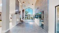 This contemporary single-floor clubhouse, branded as "Gateway," offers a striking first impression through its grand entrance lobby. The space is defined by dramatically soaring vaulted ceilings and an expansive gabled glass window above the entryway, allowing abundant natural light to pour in and frame views of the surrounding greenery. Large-format polished grey tiles extend across the main lobby floor, complementing floor-to-ceiling grey stone-clad accent walls that lend a refined, upscale atmosphere. A sleek, modern fireplace and sculptural pendant light fixtures add warmth and visual interest to the reception area.

Adjacent to the lobby, a glass-enclosed room offers a private and transparent spatial division, while a dedicated lounge area features light-toned tile flooring, vaulted ceilings with a skylight, and contemporary furnishings including curved and sectional sofas accented by abstract wall art. A billiards table anchors the games area, which is bathed in natural light from large panoramic windows. A compact kitchenette with a marble-topped counter, bar seating, pendant lighting, and light wood flooring completes the amenity spaces. Throughout, thoughtfully placed indoor plants and modern wall sconces contribute to a welcoming, sophisticated ambiance.