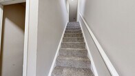 This multi-level residence is organized across two floors with an additional basement level, providing a practical and functional layout. The main living floor hosts a combined kitchen and living room area, where neutral-toned carpet extends throughout the spacious living space, which benefits from a window and sliding door that allow natural light to filter in. The kitchen features white shaker-style cabinetry, granite-look countertops, a stainless sink, and a black dishwasher, with a pass-through window connecting it to the living area.

The upper floor contains two bedrooms, including a generously sized primary bedroom, both finished with soft carpet and neutral walls. A dedicated closet provides storage, and two full bathrooms serve the residence. Each bathroom is finished with wood-look vinyl flooring, white vanities with solid countertops, large mirrors, and chrome fixtures, maintaining a clean, cohesive aesthetic throughout.

Carpeted staircases with white trim connect the levels, while the basement presents a concrete-walled utility and storage space. Overall, the home offers a straightforward, clean design with consistent neutral finishes.