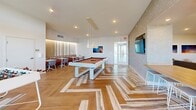 Located at 2500 SW 22nd Terrace in Fort Lauderdale, Florida, this thoughtfully designed amenity space spans a generous single floor, offering residents a well-rounded environment for relaxation and entertainment. The layout encompasses two distinct living areas and a dedicated game room, each finished with a cohesive modern aesthetic.

The first living area serves as a comfortable lounge, featuring a built-in wood-paneled media wall with open shelving, a large flat-screen TV, and warm neutral furnishings including plush seating and accent tables. Colorful artwork adorns the walls, adding a curated, residential feel to the space.

The game room is a standout feature, anchored by a full-size pool table beneath a cluster of modern pendant lights. A striking chevron-patterned hardwood floor adds visual depth, while a foosball table, booth-style seating with warm wood accents, and ambient LED lighting complete the upscale, social atmosphere.

The second living area transitions seamlessly to a covered outdoor terrace, where light wood-toned flooring extends the interior palette outside. The terrace is furnished with contemporary round tables and mixed seating, all set against an open railing that reveals expansive views of the Fort Lauderdale skyline and lush tree canopy below. Natural light floods both the interior and exterior spaces, creating a bright and airy ambiance throughout.