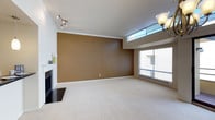 This single-floor space presents a thoughtfully designed interior that balances warmth and openness. The main living area is a standout, featuring a sleek fireplace with a dark tile surround that serves as a natural focal point. A rich, earthy brown accent wall adds depth and warmth to the room, complementing the clean white crown molding and baseboards that frame the space with a polished finish. Soft, plush carpeting covers the floor, contributing to the comfortable and inviting atmosphere. Natural light floods the room through a large sliding glass window and a high transom-style clerestory window positioned above, drawing the eye upward and enhancing the sense of vertical space. The elevated windows also provide privacy while still allowing generous daylight to filter through. Overhead, a stylish multi-arm track light fixture and a classic chandelier provide layered lighting options to suit various moods and needs. The open layout offers flexible furnishing possibilities, making it well-suited for both everyday living and entertaining. Overall, the space strikes a balance between modern design elements and a warm, welcoming aesthetic.