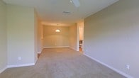 This single-floor space presents a clean, open layout designed for comfortable, everyday living. The main living area is finished with wall-to-wall carpeting in a neutral beige tone, creating a warm and cohesive atmosphere throughout. Smooth, light-colored walls with crisp white baseboards contribute to the bright and airy feel of the space.

Overhead, a ceiling fan ensures year-round comfort, while a pendant light fixture anchors the dining or secondary area, providing both functional and ambient illumination. A centrally located ceiling vent indicates a well-integrated HVAC system for climate control.

The open floor plan allows for flexible furniture arrangements and a natural flow between living zones. Visible from the main area, an adjoining room — likely a bathroom — features tile flooring and standard fixtures, offering a practical complement to the main living space.

Overall, the space is characterized by its understated, neutral palette, practical finishes, and straightforward layout, making it a versatile and functional environment well-suited for a variety of uses.