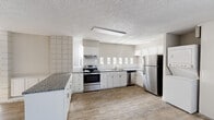 This single-floor unit offers a functional and efficient layout, comprising one bedroom, a kitchen, and a living room. The kitchen is a standout feature, equipped with white shaker cabinetry, granite countertops, a classic subway tile backsplash, and a full suite of stainless steel appliances including a range, dishwasher, and refrigerator. A stacked washer and dryer unit is conveniently housed within the kitchen area. Wood-look flooring runs throughout the kitchen and living areas, adding warmth to the neutral, light-toned interior. The living room is open and airy, benefiting from natural light filtering through vertical blinds that cover a large sliding glass door. A tiled accent wall adds a distinctive architectural detail at the junction of the kitchen and living areas. The spacious primary bedroom features plush carpeting, a generously sized sliding-door closet, and painted cinder block walls that lend the room a solid, durable character. Throughout the unit, textured ceilings and clean white trim provide a cohesive, well-maintained aesthetic. The overall ambiance is bright, neutral, and move-in ready, offering a practical living space with thoughtful finishes.