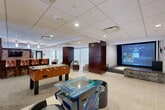 This expansive amenity and recreation center offers an impressive range of sports and entertainment facilities across a generously sized single floor. The space is divided into several distinct zones, each designed to serve a specific recreational purpose.

The game room area features a drop-ceiling with recessed lighting, rich dark-wood column accents, and light tile flooring. It is equipped with a foosball table, an arcade gaming station, and a large projection screen with a stone-accented base, ideal for gaming or media viewing. Adjacent seating areas provide comfortable spots to relax between activities.

A dedicated ping-pong room with patterned carpet flooring and large windows offers natural light and views of greenery outside. Dark wood wall paneling and a mounted flat-screen TV complete the polished look of this space.

The racquetball court features pristine white walls, hardwood flooring with regulation markings, and glass panel walls for spectator viewing. Nearby, a full indoor pickleball court impresses with soaring ceilings, a slatted white ceiling treatment, exposed brick walls, and an upper-level mezzanine overlooking the court. Vivid sports-themed artwork lines the walls, and high-top seating areas are available courtside. A bold staircase with red railings connects to the upper level, with large windows filling the space with natural light.