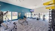 The Cypress Fitness Center at 10015 N Owasso Expressway in Owasso, Oklahoma, is a thoughtfully designed exercise facility offering a comprehensive workout experience within a single, generously proportioned room. The space features soaring ceilings accented by recessed lighting and ceiling fans, creating an open, airy atmosphere that enhances comfort during exercise.

The interior is defined by a bold teal accent wall contrasted against crisp white walls and trim, giving the room a modern, energizing aesthetic. Patterned carpet in coordinating tones of blue and gray covers the floor throughout, adding visual interest while providing a comfortable underfoot surface.

Large windows along multiple walls flood the space with abundant natural light and offer views of the outdoor pool area and surrounding grounds, creating a seamless connection to the exterior environment. A wall-mounted flat-screen television provides entertainment during workouts.

The fitness center is well-stocked with a diverse range of equipment. Cardio offerings include treadmills, elliptical trainers, and stationary bikes, while strength training is supported by a multi-station cable machine, adjustable weight benches, a dumbbell rack, kettlebells, and medicine balls. Full-length mirrors on one wall enhance the sense of space and assist with form. A small amenity station near the entrance provides towels and fitness accessories for added convenience.