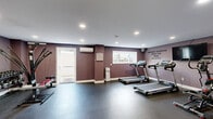 Located at 8040 Rowland Avenue in Philadelphia, PA, this fitness center and yoga studio is a thoughtfully designed wellness facility spread across a single floor. The space is divided into distinct areas, each tailored to support a specific aspect of health and fitness.

The fitness room features deep plum-colored walls, durable rubber flooring, and recessed lighting, creating an energizing atmosphere for cardio and strength training. Treadmills, a rowing machine, and resistance equipment are housed here, accompanied by a wall-mounted split AC unit and motivational wall lettering that reads, "The body achieves what the mind believes."

The yoga and studio area offers a striking contrast, with light hardwood-style flooring, floor-to-ceiling glass partitions, and elegant floral wallpaper that lends a serene, welcoming ambiance. A built-in wooden shelving unit provides organized storage for books, towels, and accessories, including a dedicated cubby section for shoes. A glass-paneled door allows natural light to filter into the space.

A long corridor connects the two primary areas, with white paneled doors providing access to additional rooms and storage. The facility's thoughtful layout, calming design palette, and purpose-built amenities make it a comprehensive destination for fitness and mindfulness in the Northeast Philadelphia community.