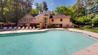 Located at 500 Harbor Pointe Pkwy in Sandy Springs, Georgia, this outdoor amenity area centers around a generously sized swimming pool with clear, turquoise water. The pool features a geometric design with gently sloping entry points and is surrounded by a wide, open deck that provides ample space for relaxation and recreation. Encircling the pool deck is an extensive collection of light-green sling lounge chairs, ideal for sunbathing, along with vibrant green patio umbrellas that offer shaded seating areas for those seeking respite from the sun. The surrounding landscape is lush and well-maintained, featuring a variety of mature trees — including towering pines — along with sculpted shrubs, ornamental plantings, and tiered garden beds that create a natural, park-like setting. A clubhouse-style building with a chimney and an elevated deck is visible at one end of the pool, adding architectural character to the space. The combination of clear open skies, dense tree canopy, and thoughtfully arranged outdoor furnishings creates a serene and resort-like atmosphere. The pool area is fully fenced for safety and privacy, making it a well-appointed outdoor retreat within the Sandy Springs community.