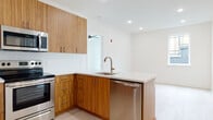 This single-floor residence showcases a thoughtfully designed, contemporary interior with a clean, modern aesthetic throughout. The open-concept kitchen and living area serves as the heart of the space, featuring striking warm-toned wood cabinetry with brushed metal hardware, a quartz countertop peninsula with an integrated sink and stainless steel dishwasher, and a herringbone tile backsplash. The kitchen is fully equipped with a full suite of stainless steel appliances, including a refrigerator, range, microwave, and dishwasher. Light hardwood flooring flows seamlessly throughout the entire unit, complementing the bright white walls and recessed lighting that keep the space feeling open and airy. The living area benefits from natural light through a window overlooking an exterior fence line. The bedrooms are generously sized, with one room featuring a large picture window and a ceiling fan, offering expansive rooftop views of the surrounding neighborhood. A second bedroom includes its own window with natural light. The unit includes two bathrooms: one finished with large-format marble-look tile on both the floors and shower walls with a frameless glass enclosure and floating vanity, and a second bathroom featuring classic white subway tile surrounding a soaking tub and a modern floating vanity.